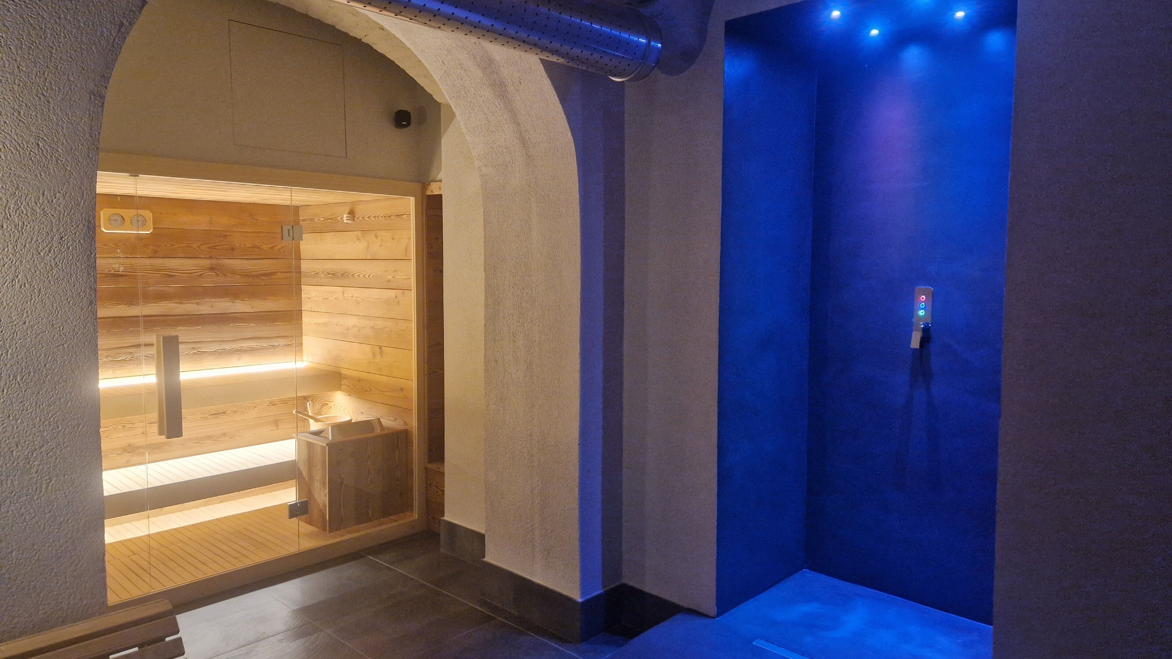 Un moderno espacio de spa con una pared de madera, una sauna con paredes de vidrio y una habitación iluminada en azul.