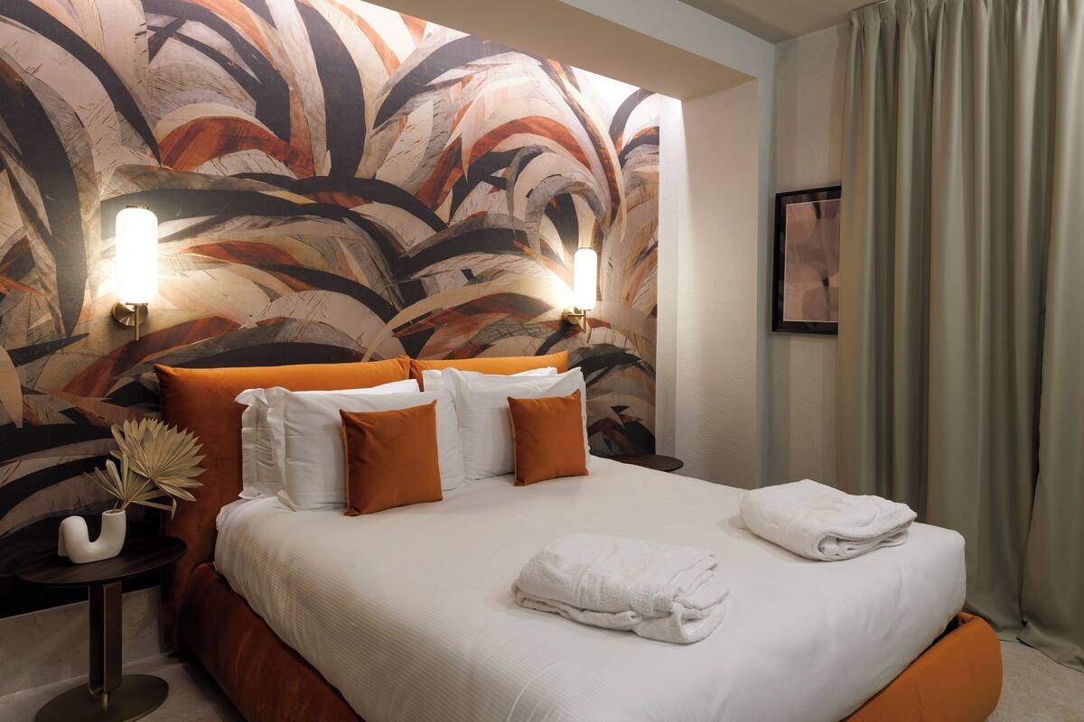 Habitación de hotel con un amplio mural abstracto en la pared, una cama con ropa de cama blanca y naranja, dos lámparas de noche y una cama bien hecha con toallas.