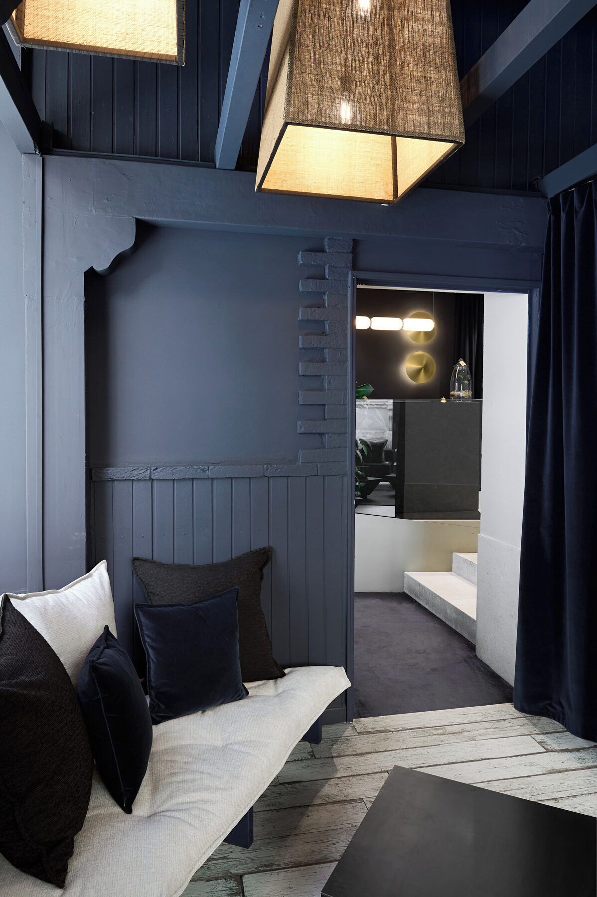 A modern interior with a dark blue wall, a wooden bench with cushions, a hanging light fixture, and a staircase leading to another level.