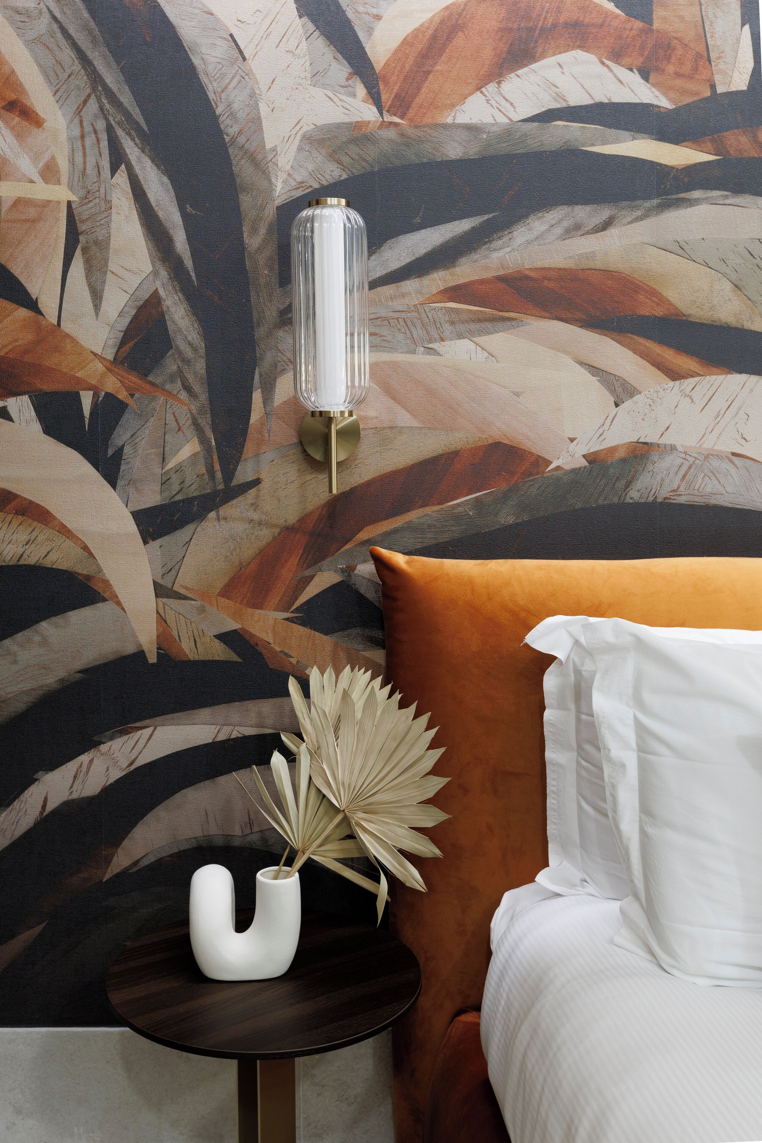 A bedroom with a bed, a nightstand, a wall-mounted lamp, and a decorative vase with dried plants.