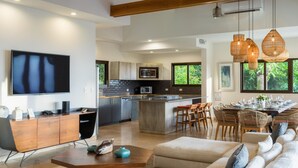 Une cuisine moderne avec un îlot central, un coin repas avec des chaises en osier, un salon avec un canapé et une table basse, et un grand téléviseur fixé au mur.