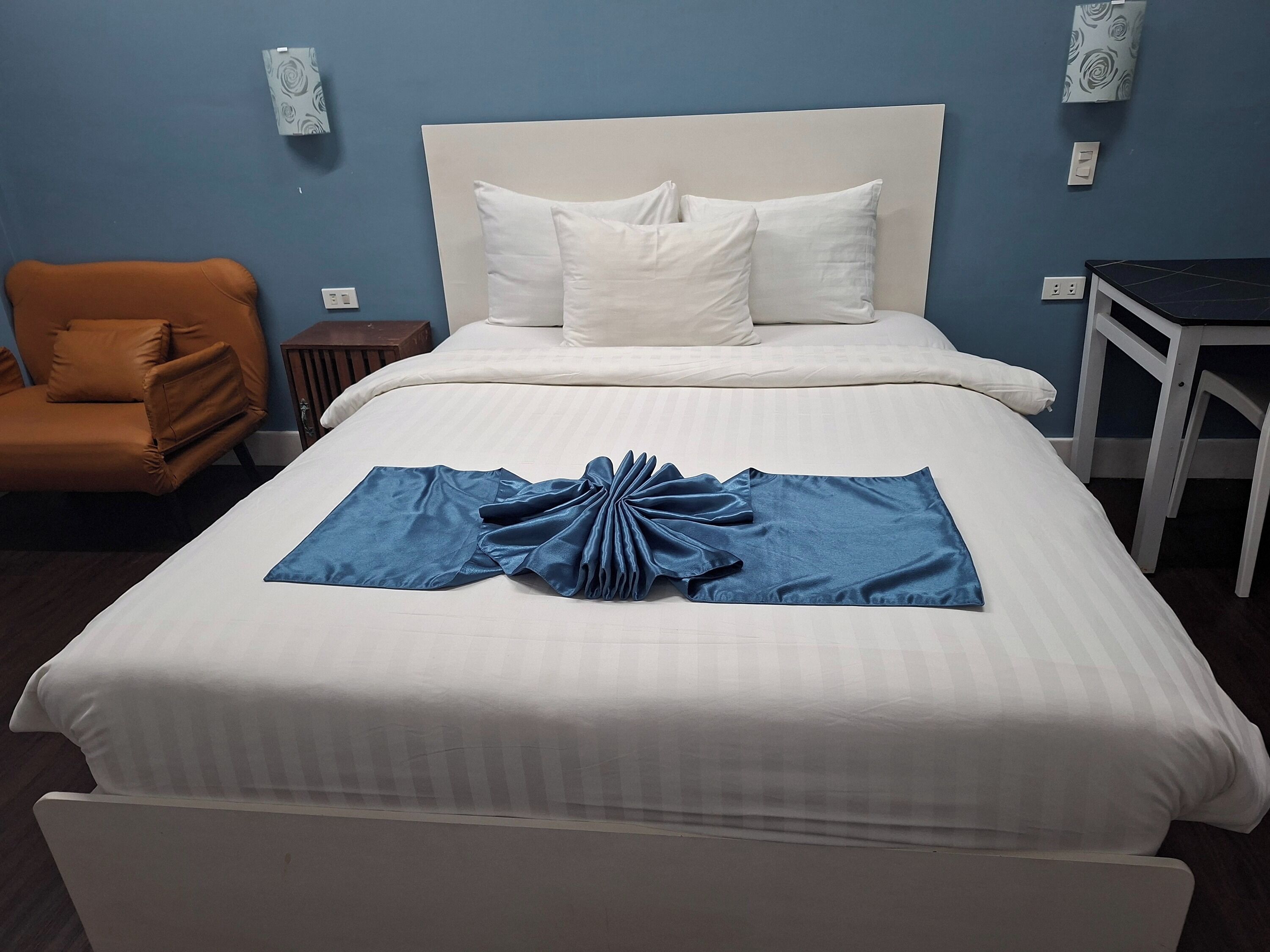 Een net opgemaakt bed met witte lakens, een blauwe handdoek en een bruine lederen fauteuil.