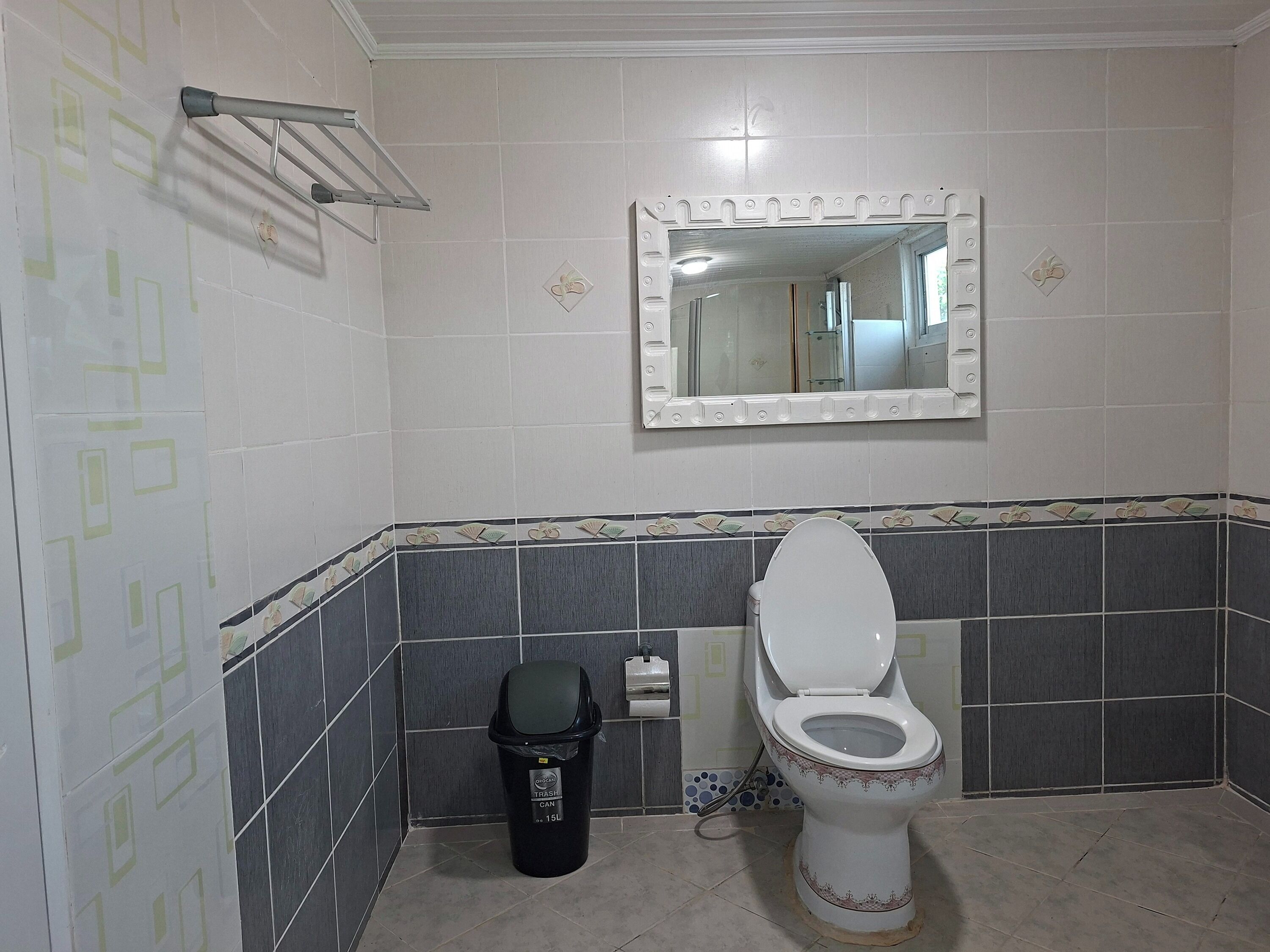 Une salle de bain avec des toilettes, une poubelle, un miroir et des murs carrelés.