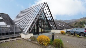 Un edificio moderno con tetto spiovente e ampie finestre vetrate, circondato da un'area pavimentata con cespugli fioriti di colore giallo e un'auto parcheggiata.