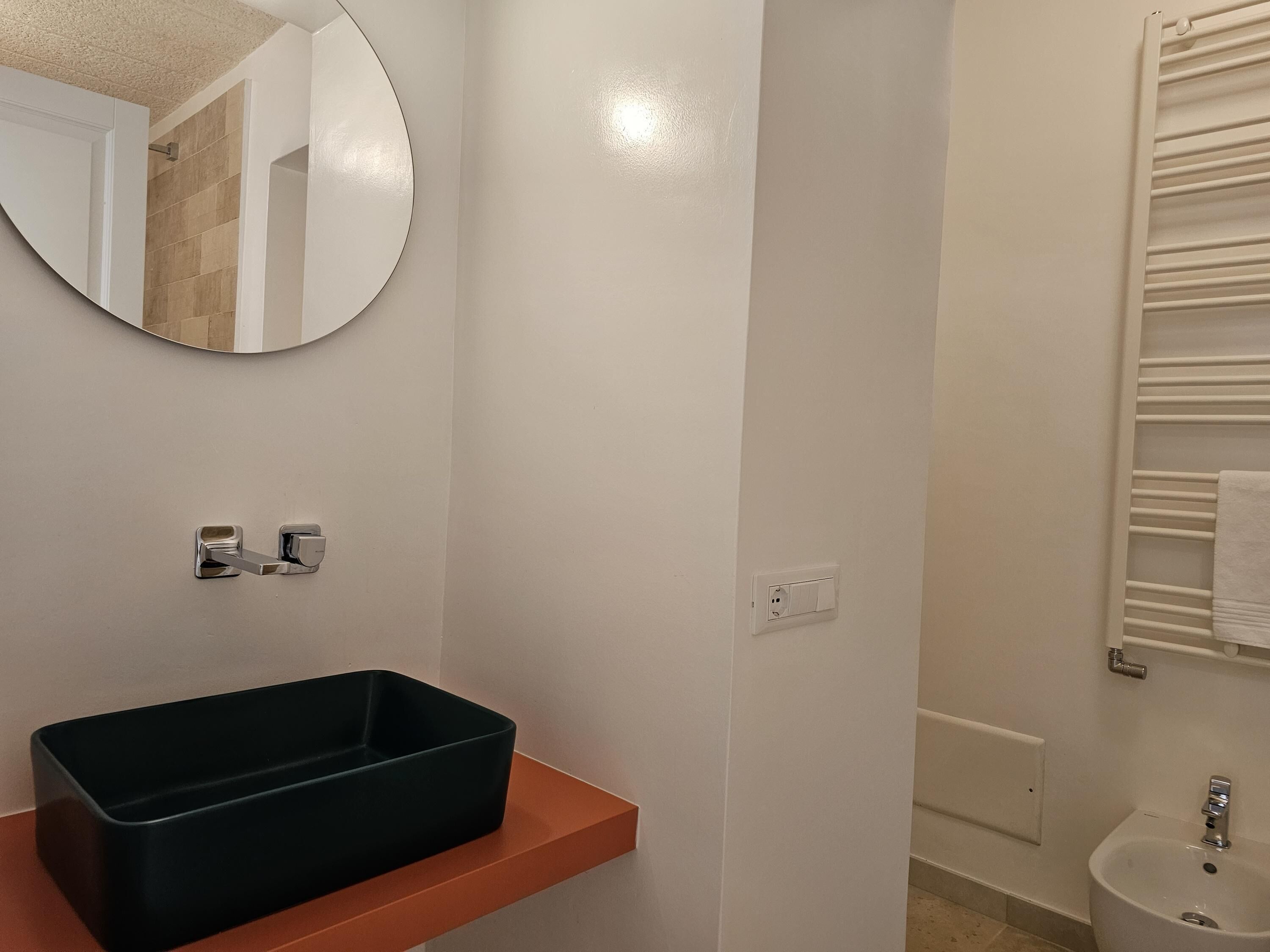 A bathroom with a rectangular black basin on an orange countertop, a round mirror, a towel rack, and a white sink.