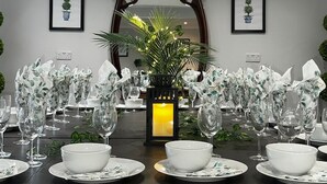 Een eettafel gedekt met witte borden, kommen en wijnglazen, omringd door potplanten en ingelijste foto's aan de muur.