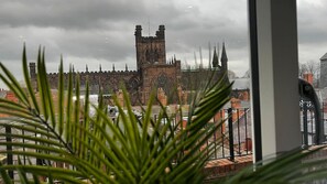 A view of a historic building with a prominent tower and spires, seen through a window with a plant in the foreground. - The Gathering On The Rows - City Centre (Chester)