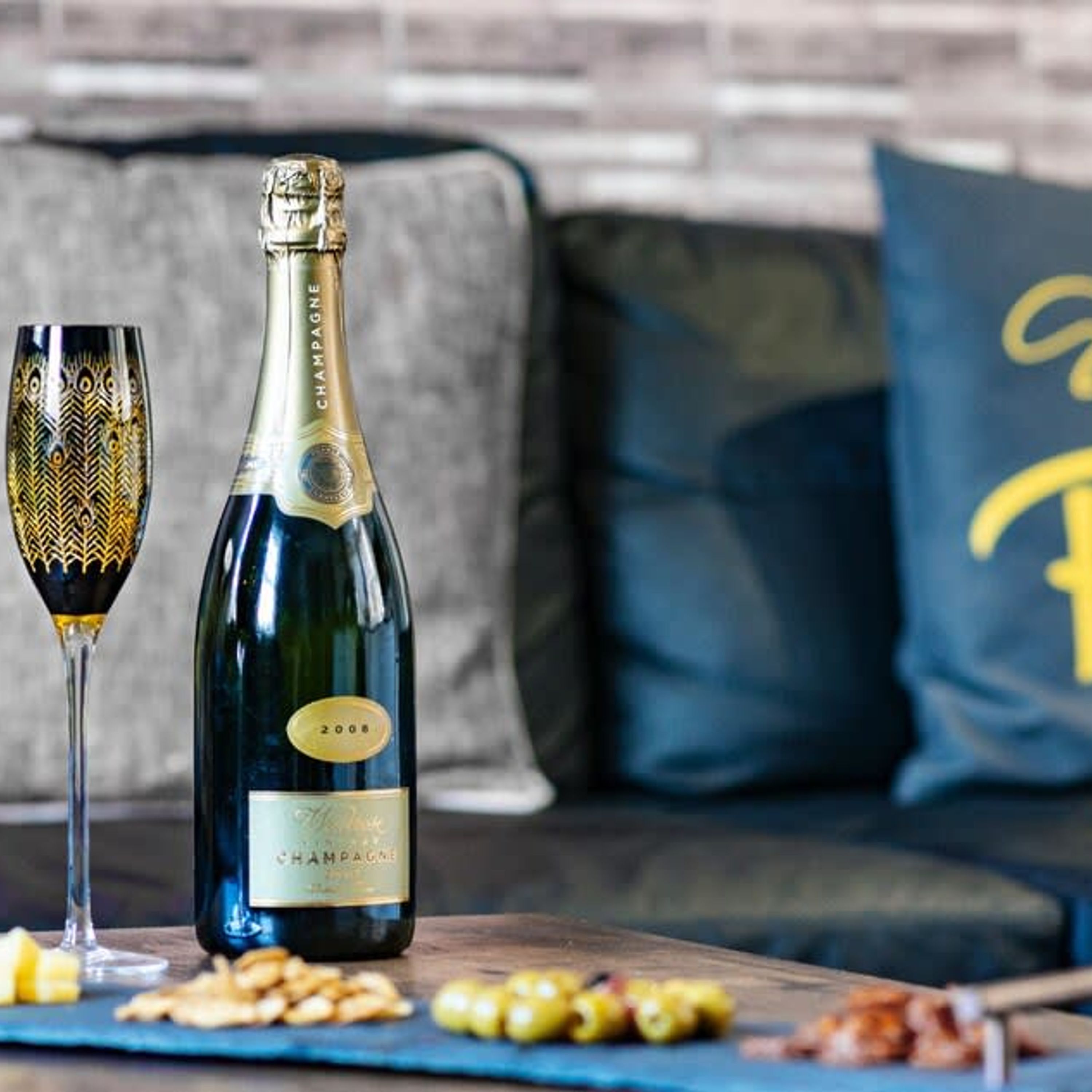 A bottle of champagne and a glass on a table with snacks, set against a modern living room backdrop.