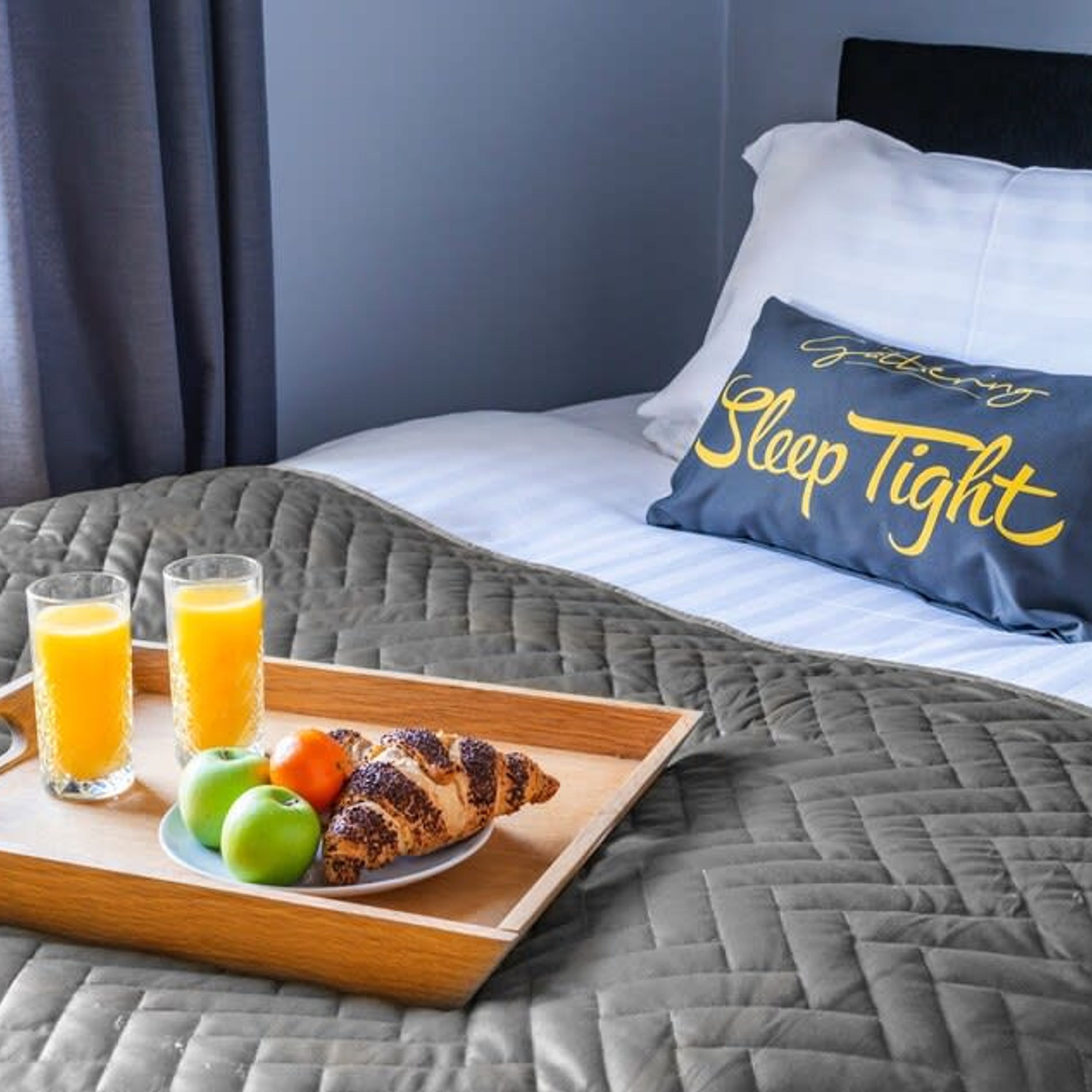 A neatly made bed with a pillow that reads "Sleep Tight" and a tray with croissants, juice, and fruit.