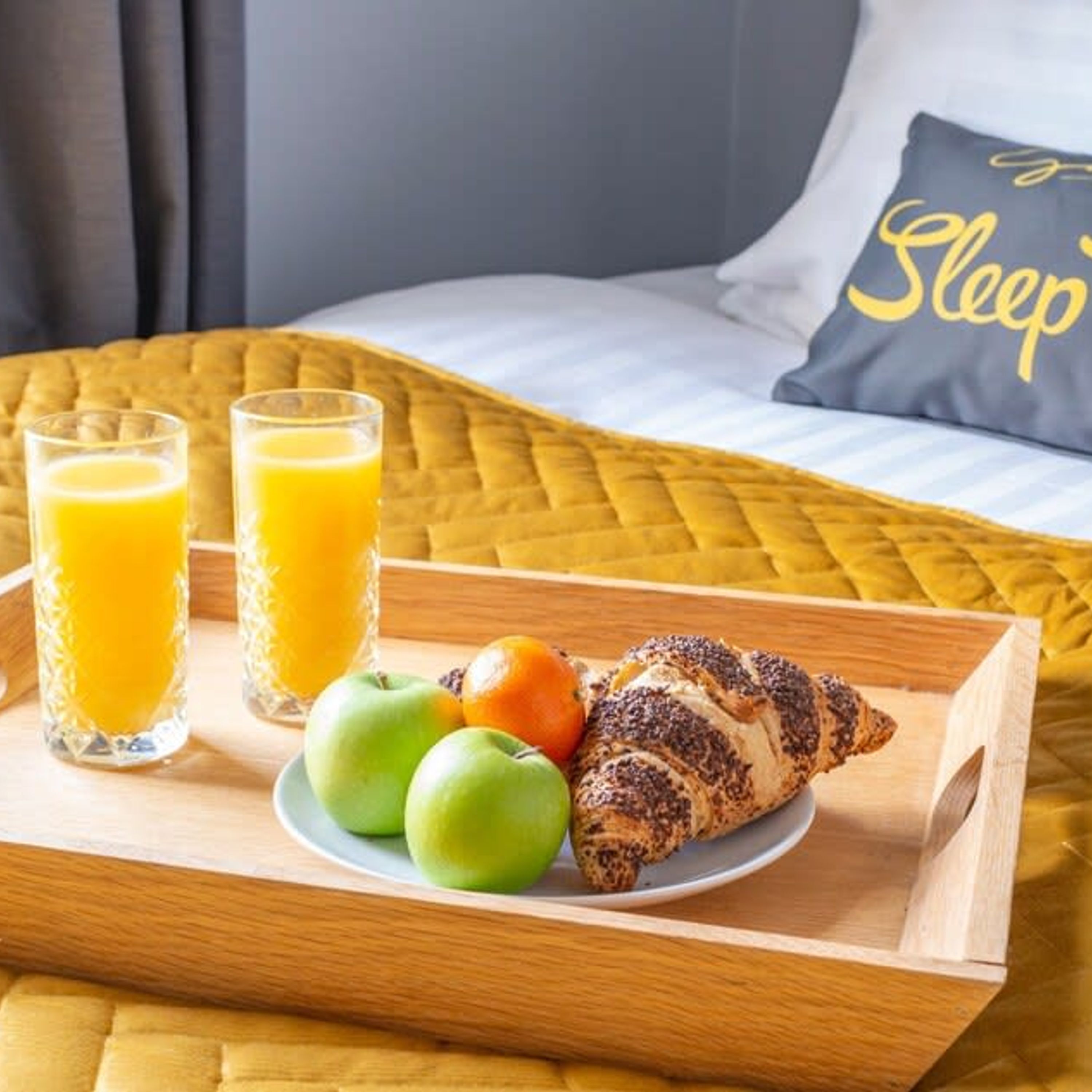 Ein Frühstücksbrett mit Croissants, Äpfeln und Orangensaft auf einem Bett mit einer gelben Bettdecke und einem Kissen mit dem Aufdruck 'Sleep'.
