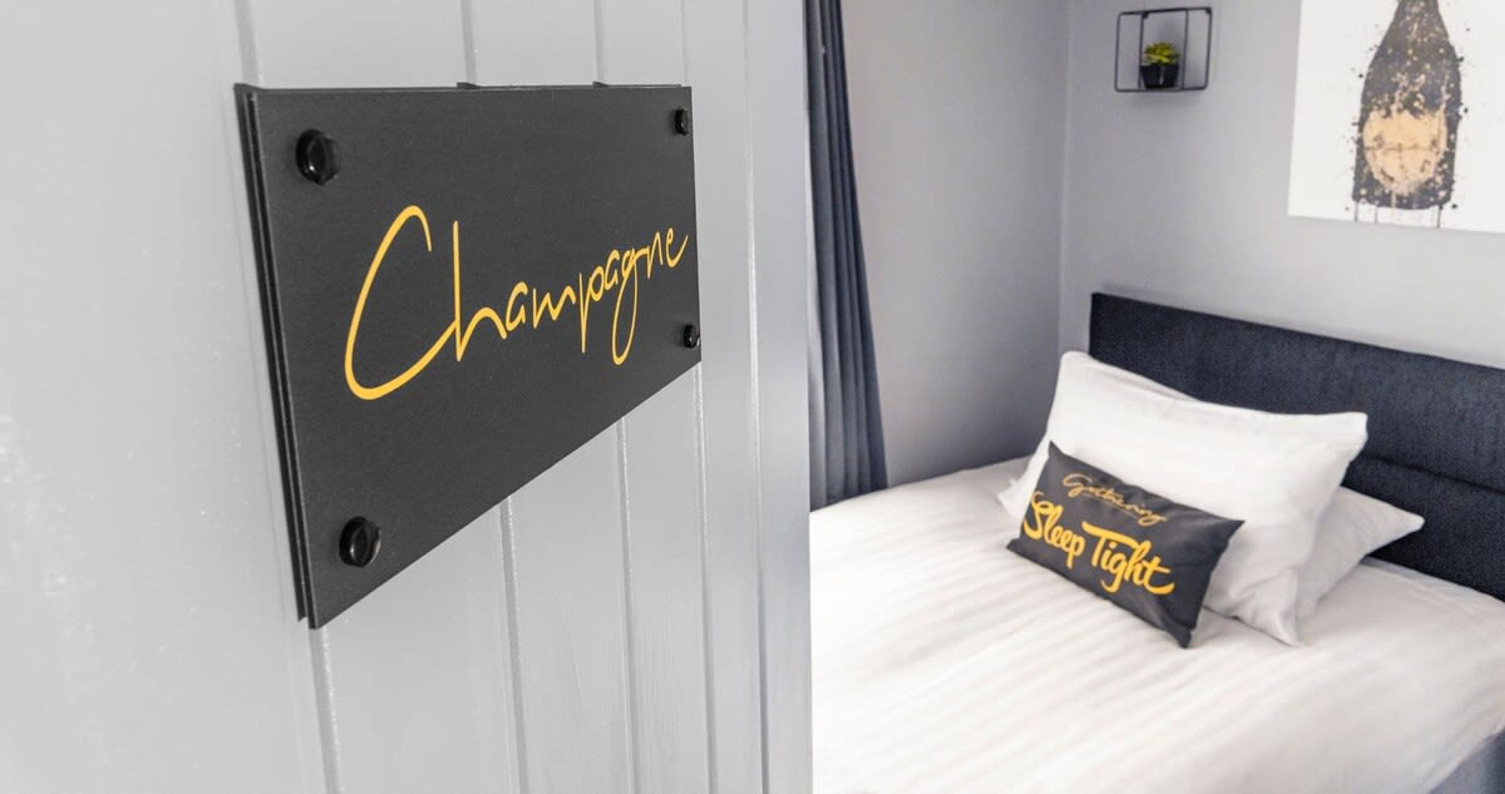Un lit avec une literie blanche et un oreiller orné de l’inscription « Bonne nuit », un panneau noir avec l’inscription « Champagne » et une petite plante posée sur une étagère.