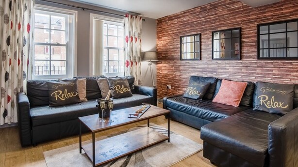 Un salon moderne avec deux canapés en cuir, chacun agrémenté de coussins «Relax», une table basse avec des boissons et un mur aux motifs de briques avec des fenêtres.