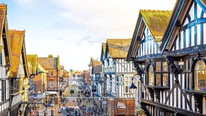 A historic street with traditional half-timbered buildings, cobblestone pavement, and a central square with a fountain. - The Lombard House - City Centre (Chester)
