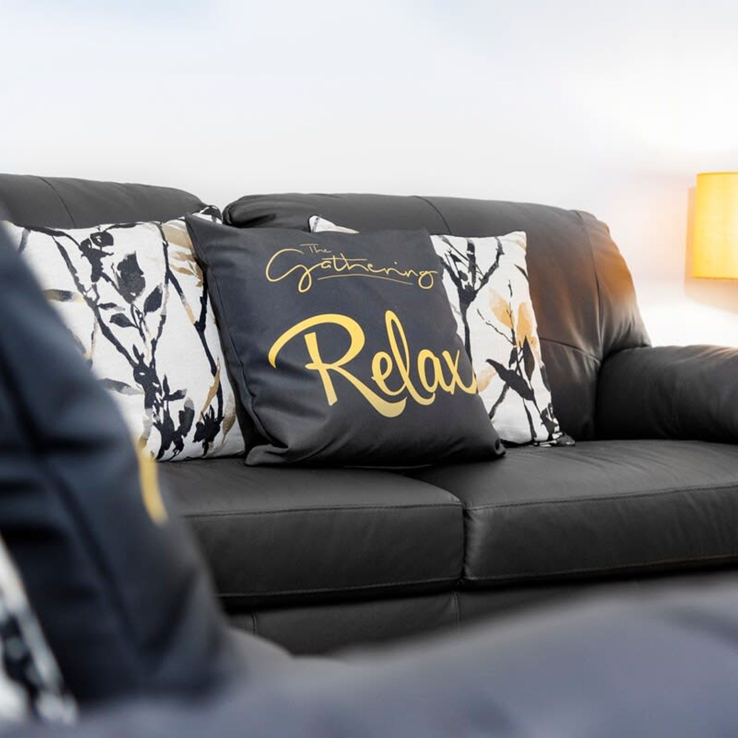 A black leather sofa with a decorative pillow featuring a floral pattern and the words "The Gathering Relax".