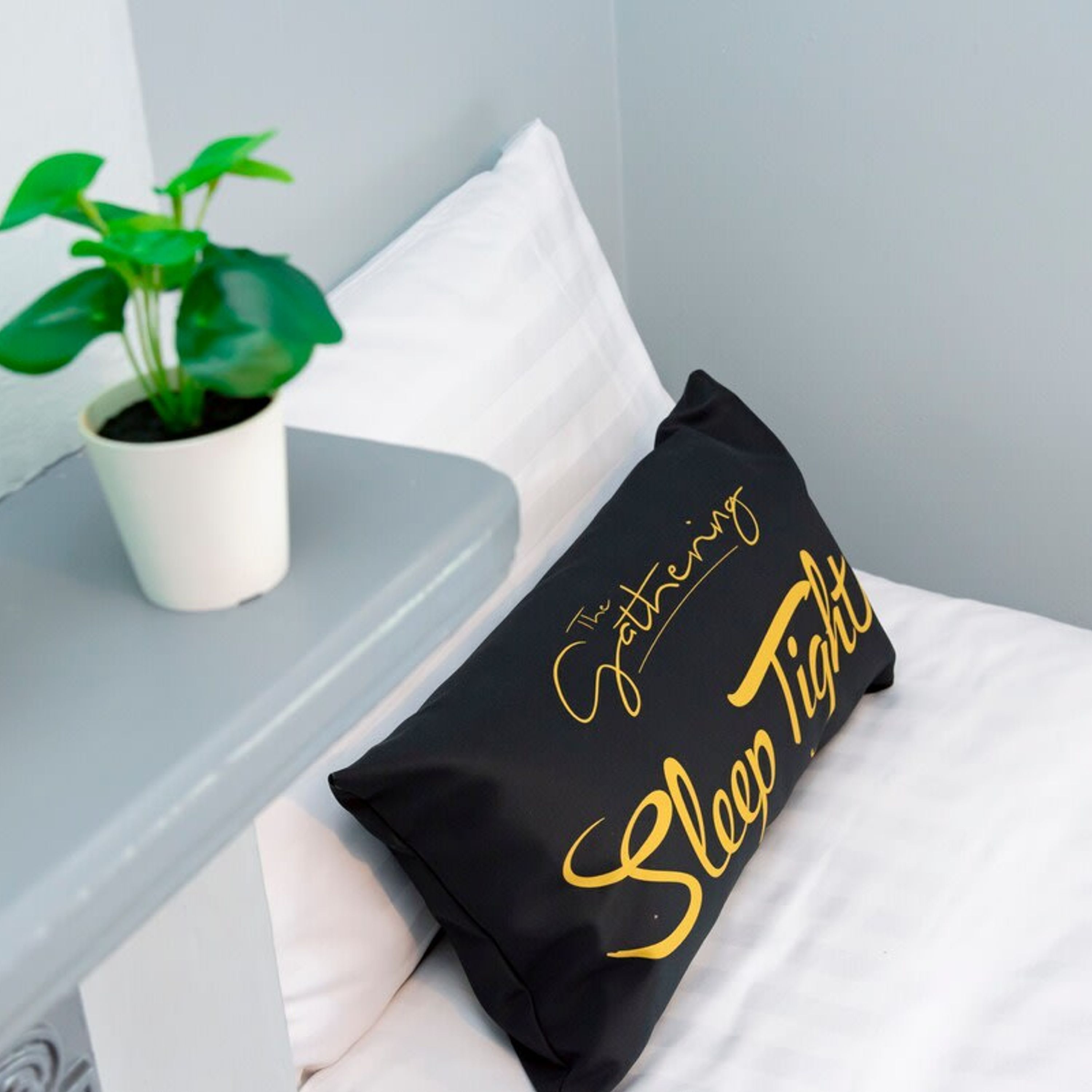 A bed with a black pillow featuring gold text, a white pillow, and a small potted plant on a white bedside table.