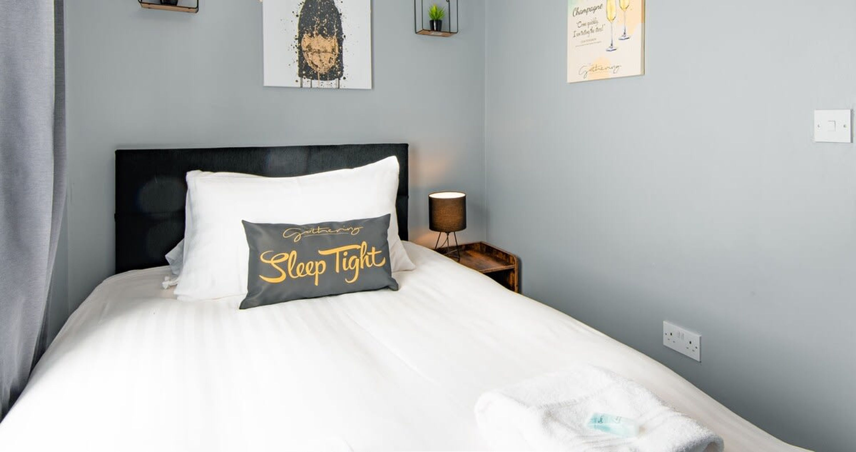 Un letto rifatto con cura, con un cuscino etichettato "Sleep Tight", in una camera d'albergo dalle pareti azzurrine, un comodino con una lampada e una piccola pianta al muro.