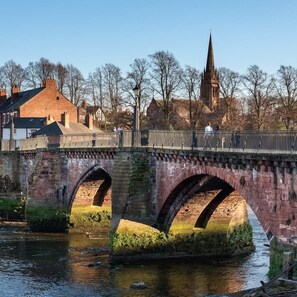 Een stenen brug met drie bogen over een rivier, leidend naar een stad met historische gebouwen en een kerktoren op de achtergrond.