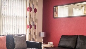 A living room with two black sofas, a wooden floor, a red wall, and a mirror. - The Racecourse Gathering 5 Bed - Sleeps 12 City Centre (Chester)
