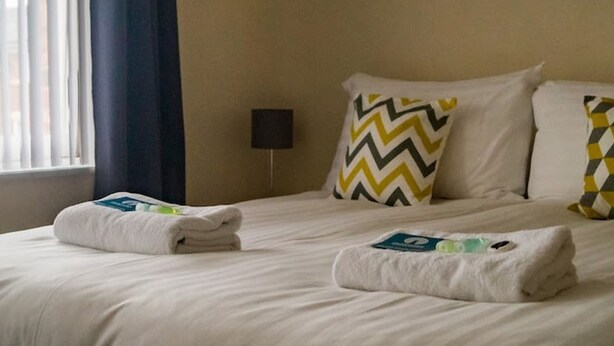 A hotel room with a bed, two pillows, a nightstand, and a window with curtains. - The Racecourse Gathering 5 Bed - Sleeps 12 City Centre (Chester)