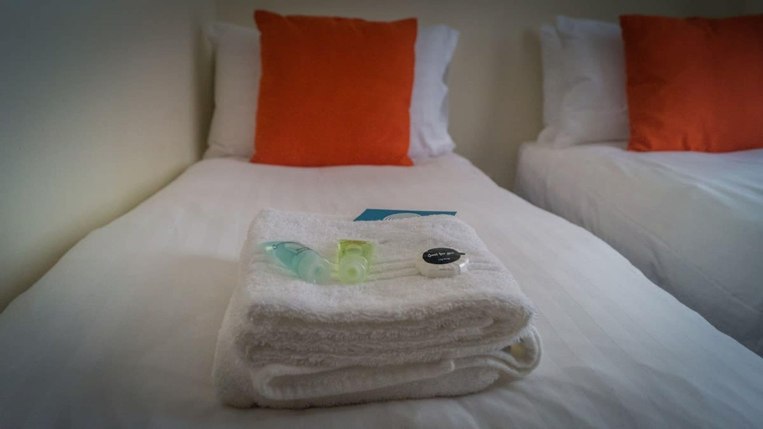 Cama bem arrumada com uma toalha branca dobrada ao centro, um cartão azul e duas garrafinhas.