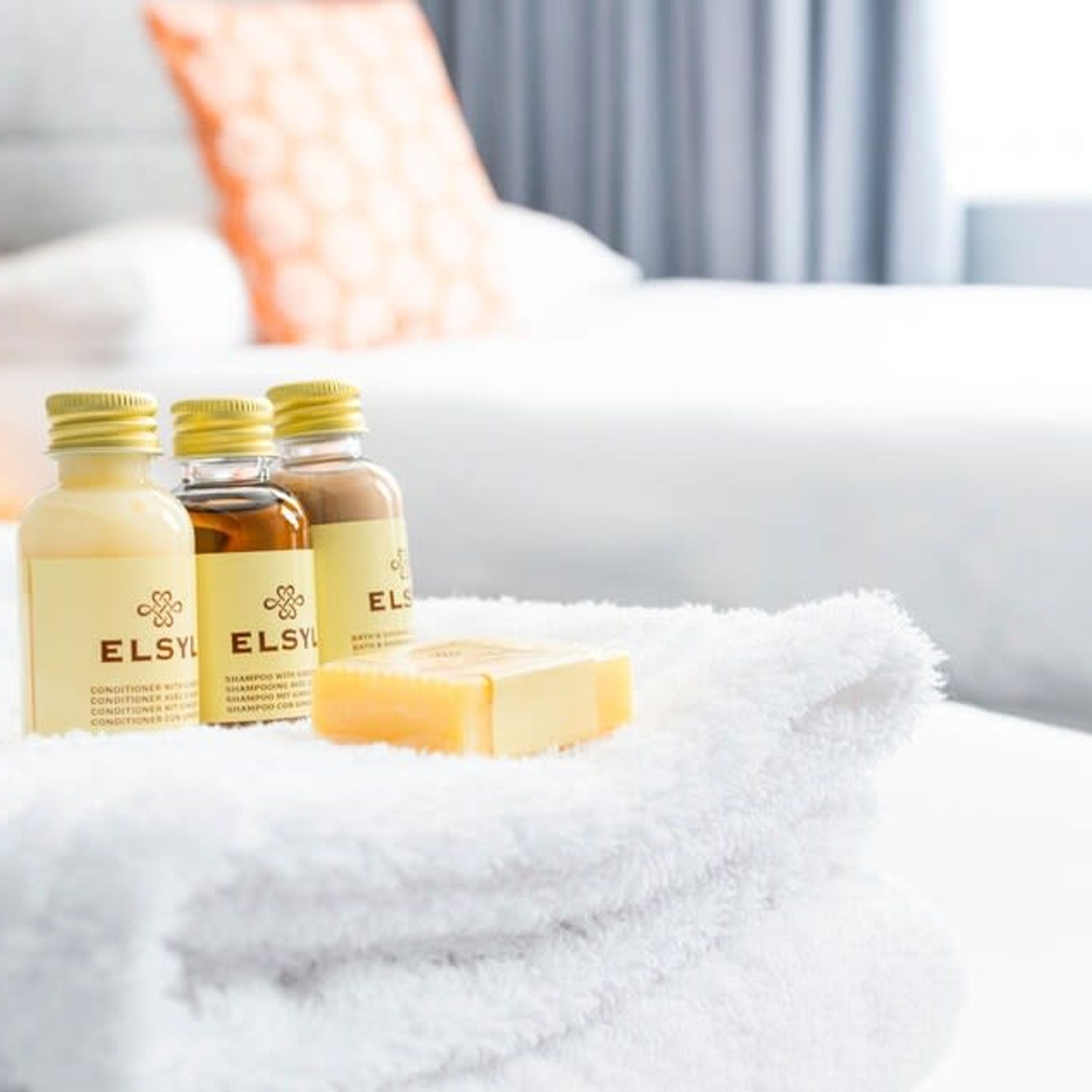 A bed with a white fluffy blanket, three bottles of ELSY shampoo, and a bar of soap on the bed.