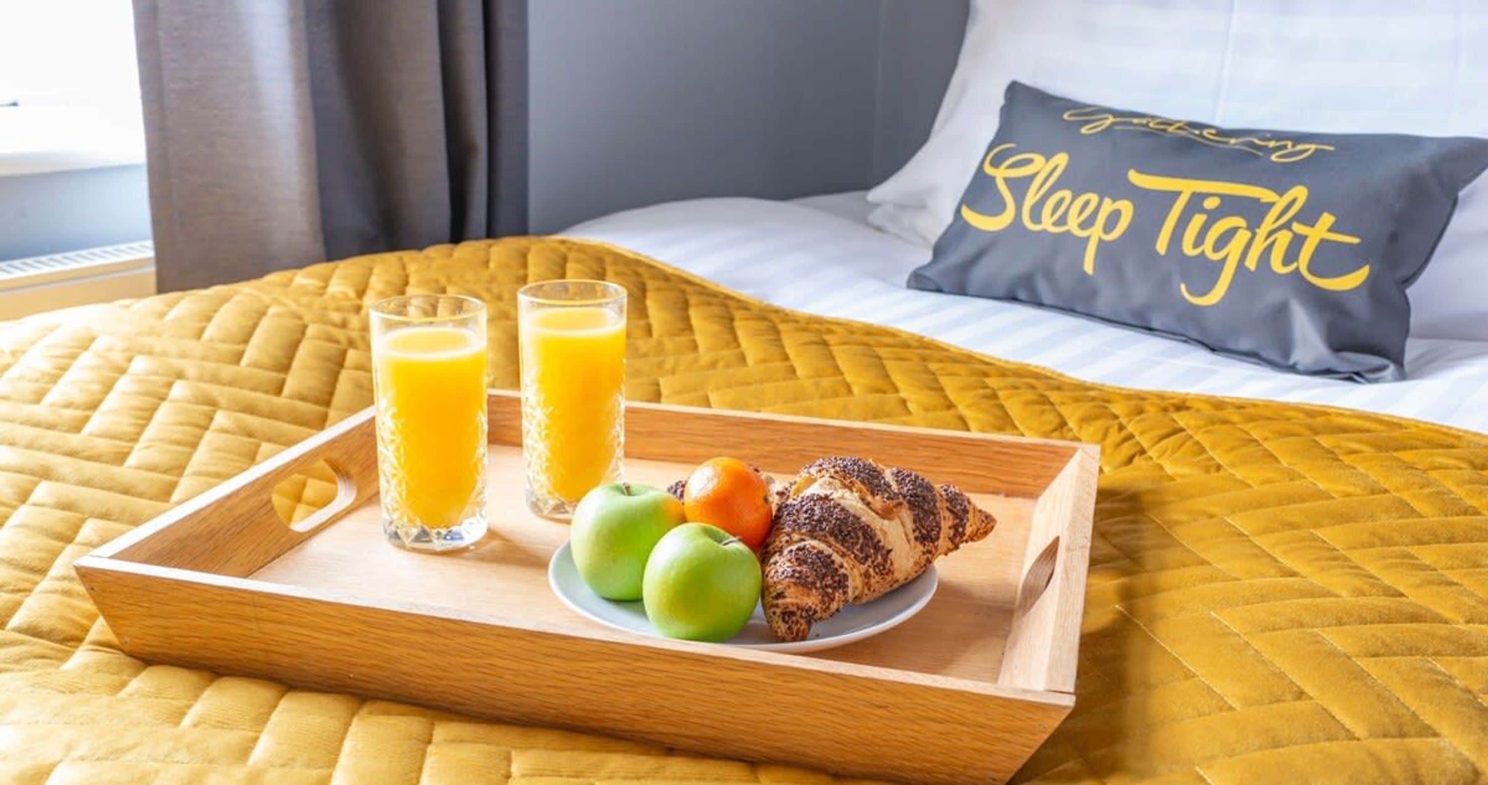 Bandeja de desayuno con vasos de jugo, manzanas y un croissant sobre una cama con una colcha acolchada amarilla y un almohadón con el texto 