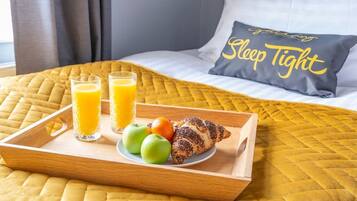 Bandeja de desayuno con vasos de jugo, manzanas y un croissant sobre una cama con una colcha acolchada amarilla y un almohadón con el texto