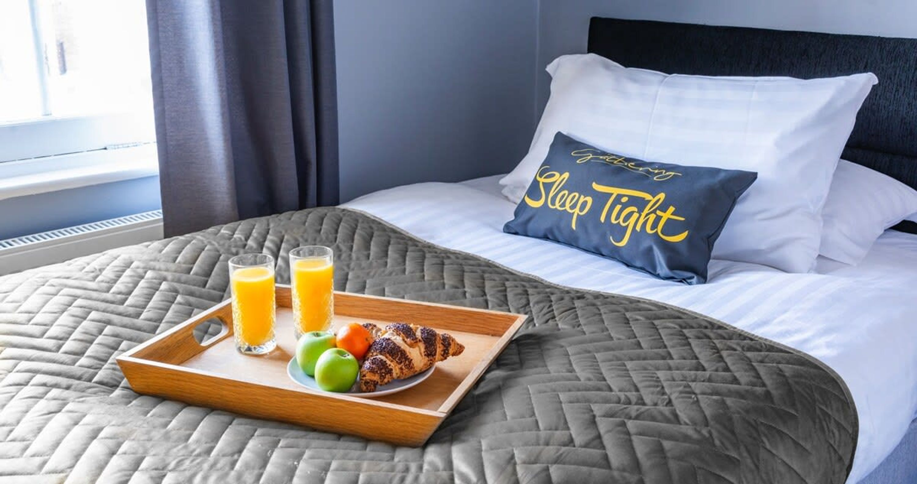 A neatly made bed with a gray quilt, a tray with orange juice and croissants, and a pillow with "Sleep Tight" text.