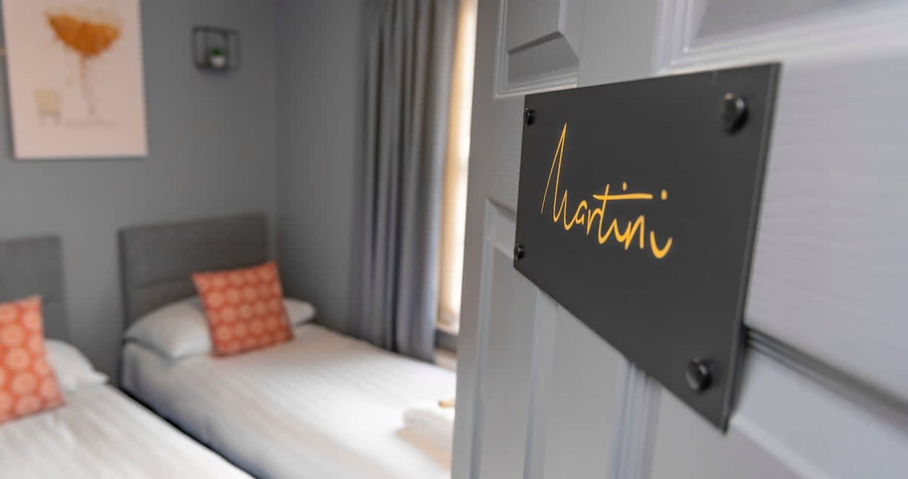 Ein Hotelzimmer mit zwei Betten, jedes mit orangefarbenen Kissen, und einem Schild an der Tür mit dem Namen Martin.