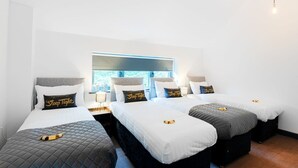 Ein Hotelzimmer mit drei Einzelbetten, jedes mit einem Kopfkissen, auf dem 'Schlaf gut' steht. Das Zimmer verfügt über ein großes Fenster mit Blick auf Bäume und einen Holzbalken an der Decke.