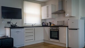 Eine kompakte Küche mit weißen Schränken, einem Geschirrspüler, einem Backofen, einem Kühlschrank und einem Fenster mit Jalousien.