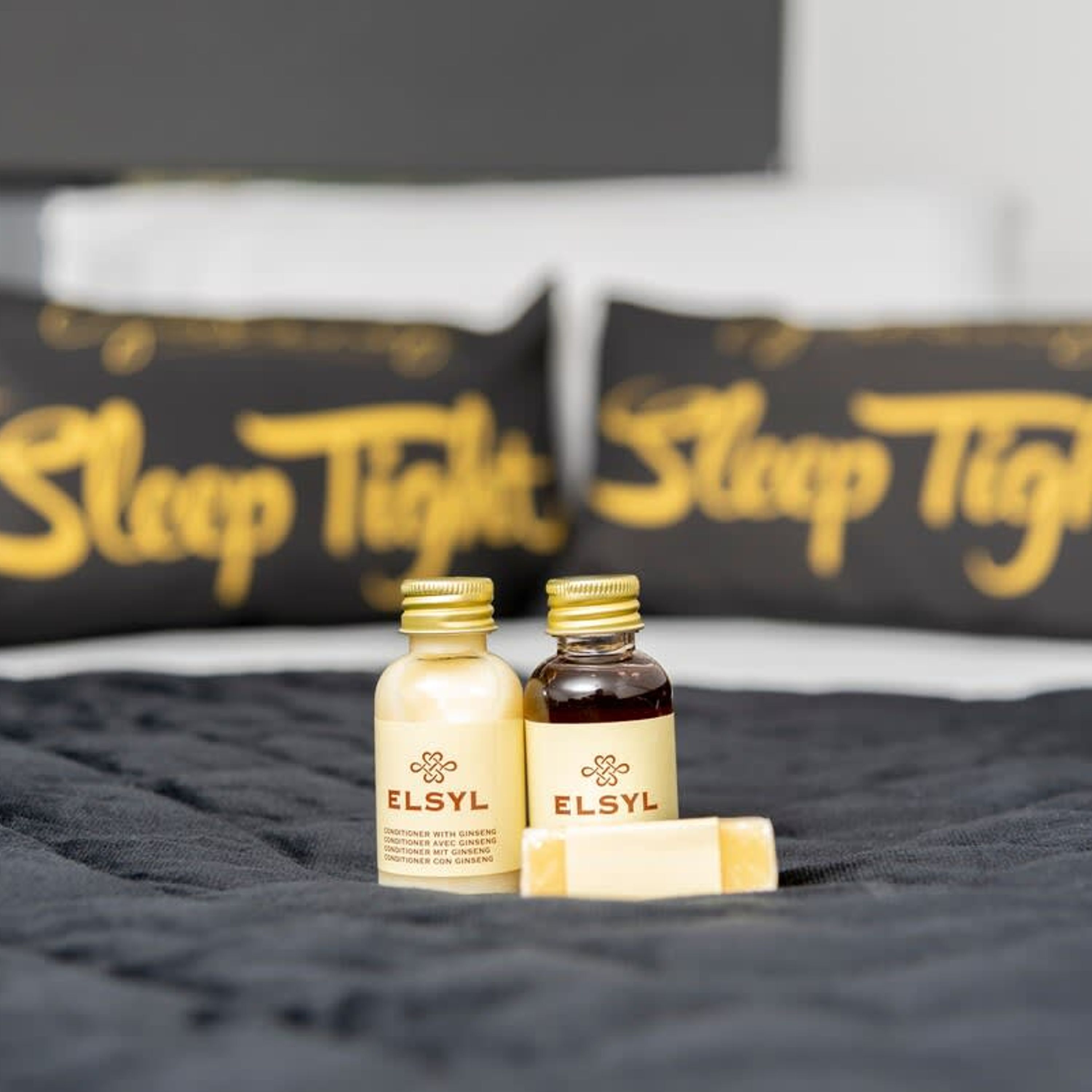 Two bottles of ELSYL conditioner with ginseng on a black bedspread, with pillows in the background that say "Sleep Tight".