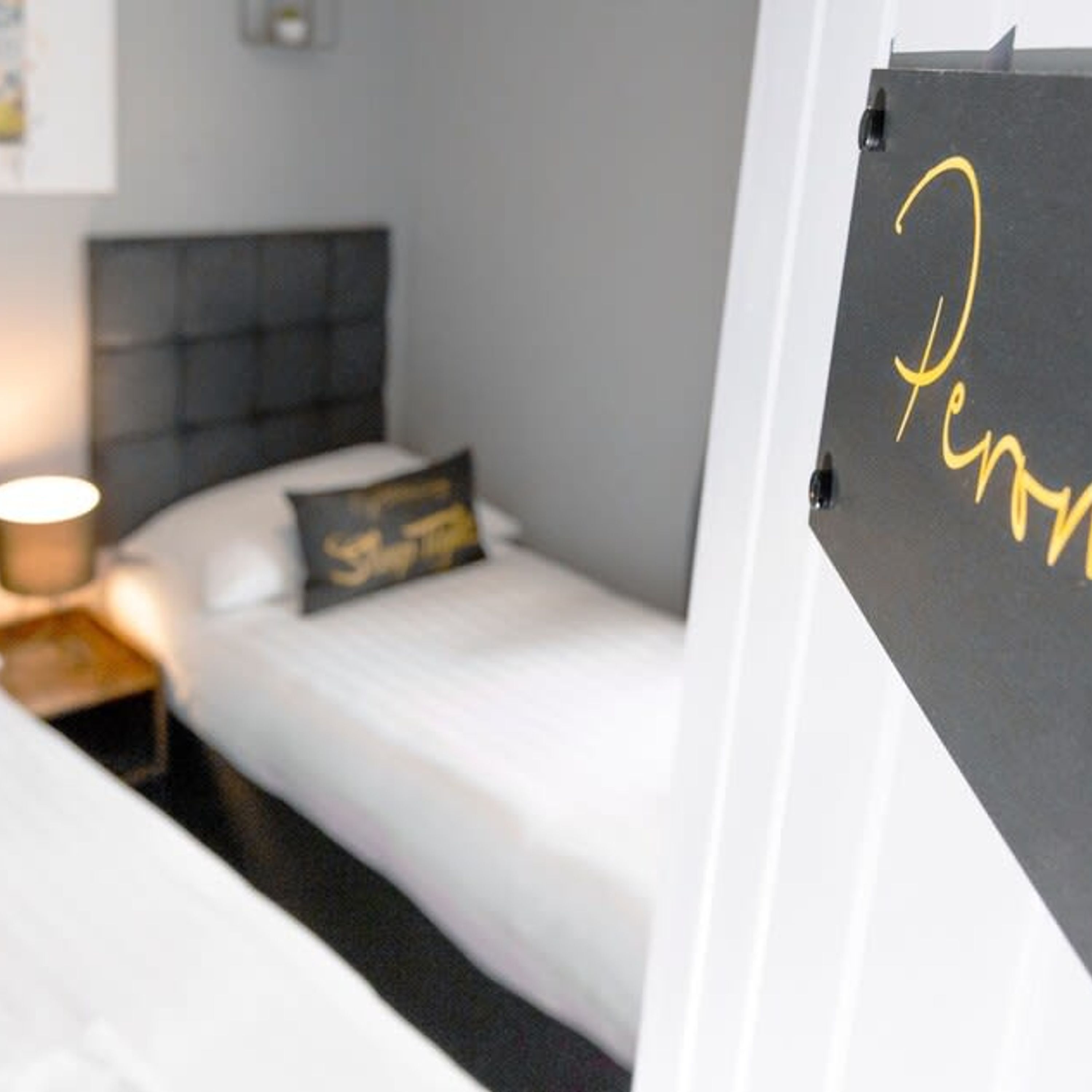 A hotel room with a bed, a nightstand, a lamp, and a blackboard with the word "Penny" written on it.