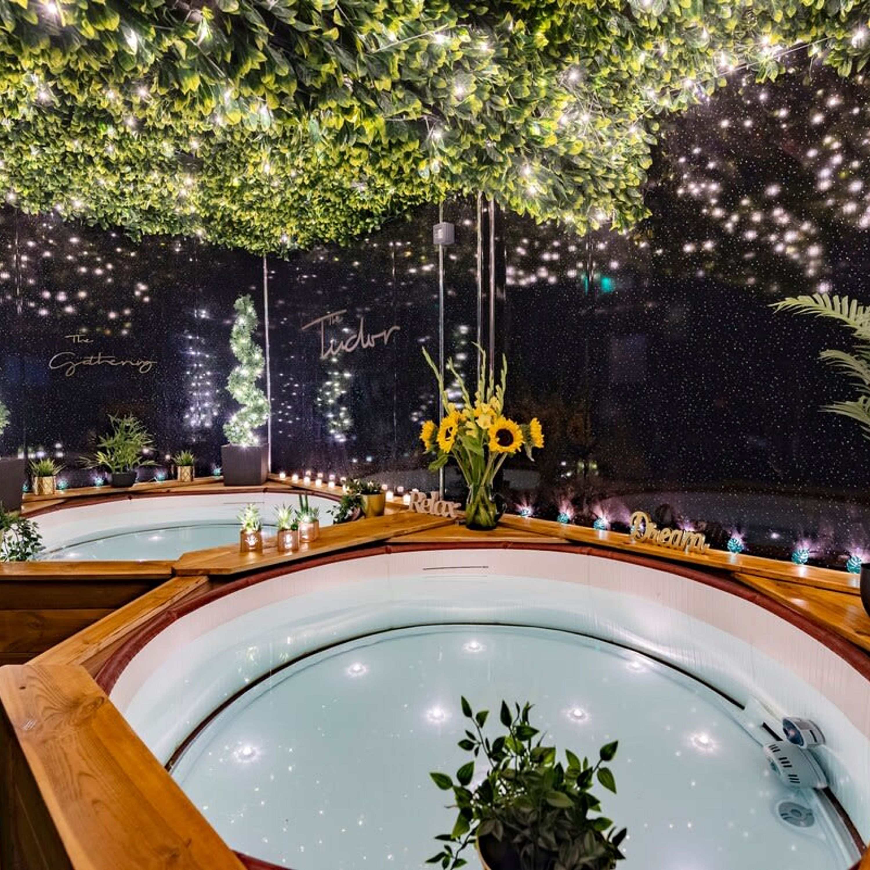 A spa area with multiple hot tubs, a ceiling adorned with greenery, and a wall with the words "The Gathering" and "Tudor".