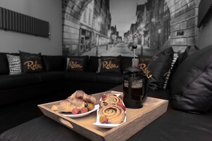 Una bandeja con pasteles y una cafetera sobre una mesa de madera en una sala de estar con un sofá de cuero negro y almohadas con la palabra 'Relax'.