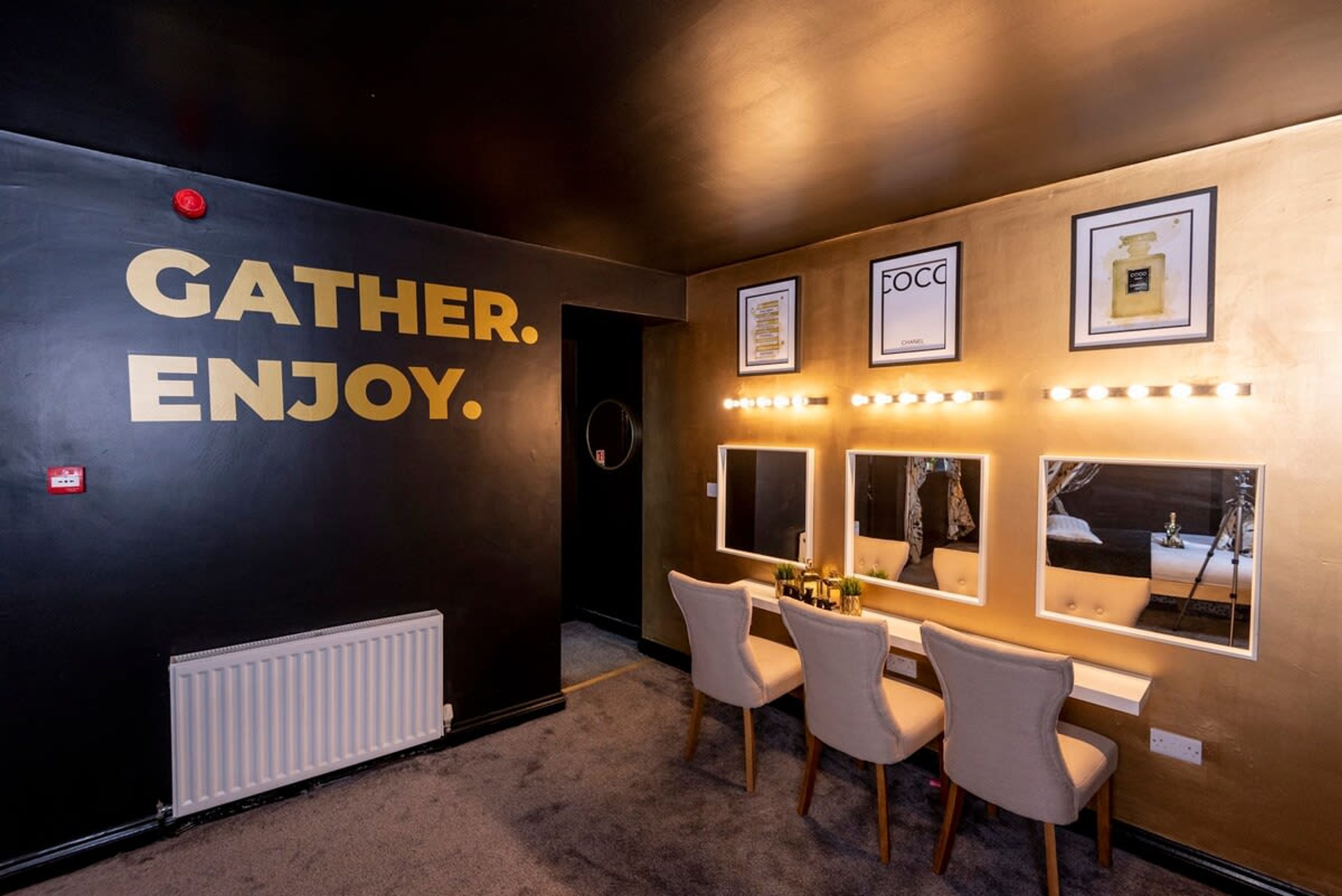 A hotel lobby with a black wall featuring the text "GATHER. ENJOY." and framed pictures of perfumes on the wall.