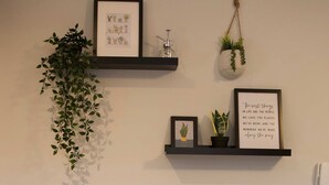 Une pièce avec un canapé gris, des coussins décoratifs, un téléphone fixé au mur et des étagères ornées de photos encadrées et de plantes.