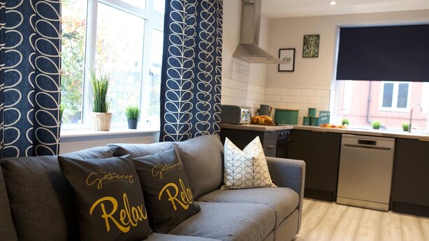 Un salon moderne avec un canapé gris, des coussins décoratifs, une fenêtre avec des rideaux et une cuisine en arrière-plan.