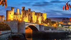 A historic castle with a stone bridge over a river, surrounded by autumn foliage. - The North Wales Gathering - 9 Bedroom - EV Charging (Flint)
