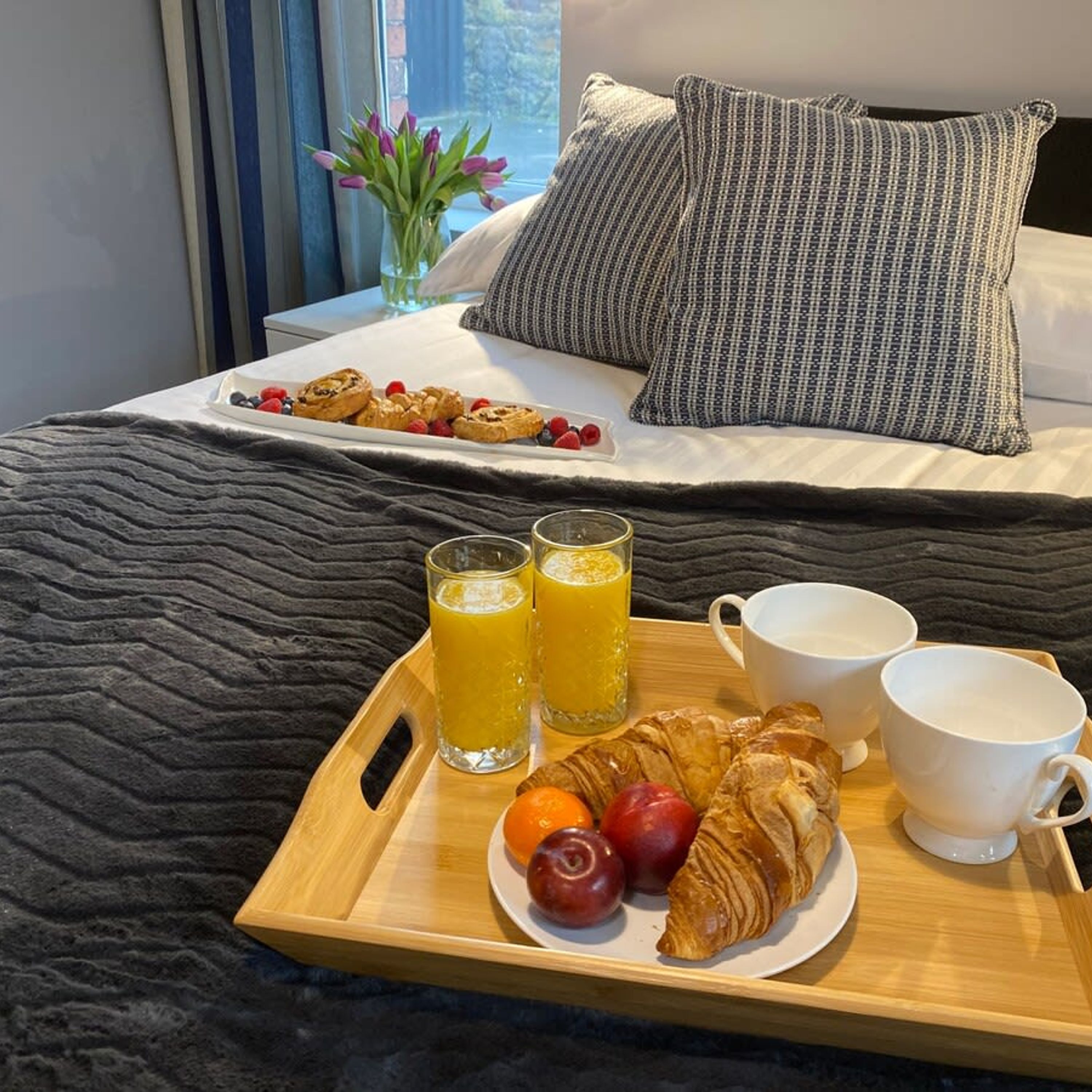 Una bandeja de desayuno ordenada con cruasanes, frutas, jugo y tazas de café sobre una cama con vistas a una ventana y un florero.