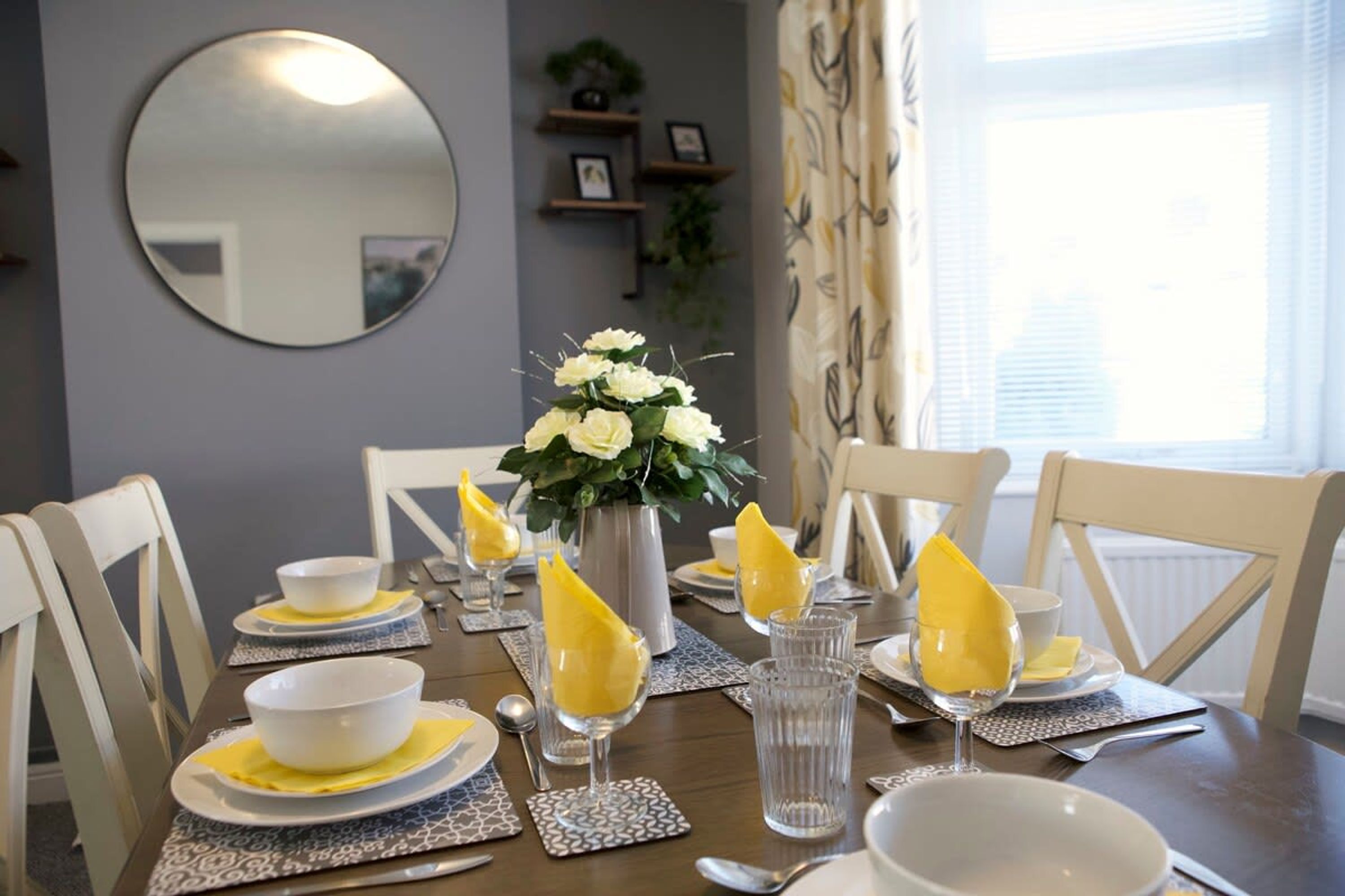 A dining table set with white plates, bowls, and glasses, accompanied by yellow napkins and a vase of white flowers.