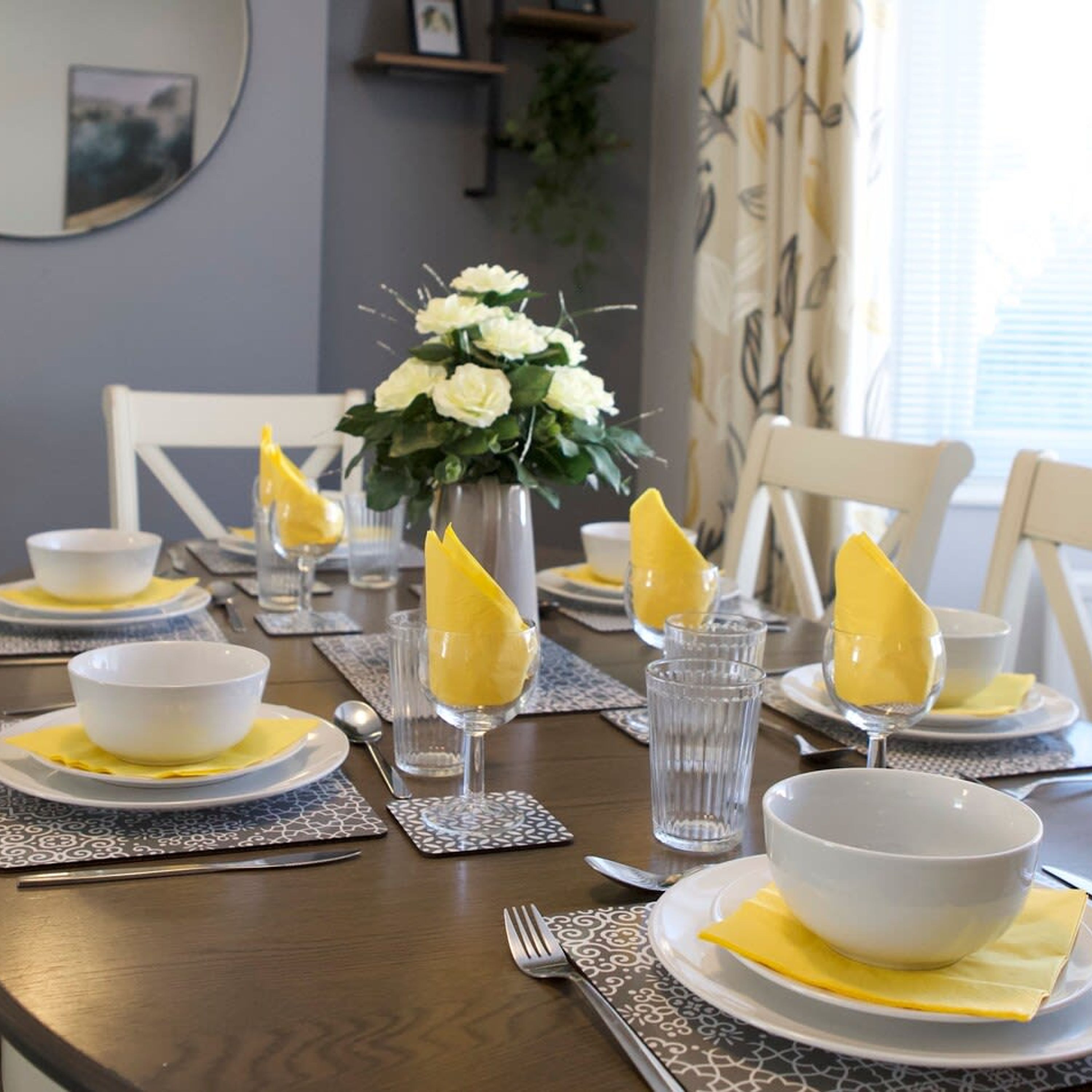 Una mesa de comedor dispuesta con cuencos blancos, servilletas amarillas y vasos transparentes, con un jarrón de flores blancas en el centro.