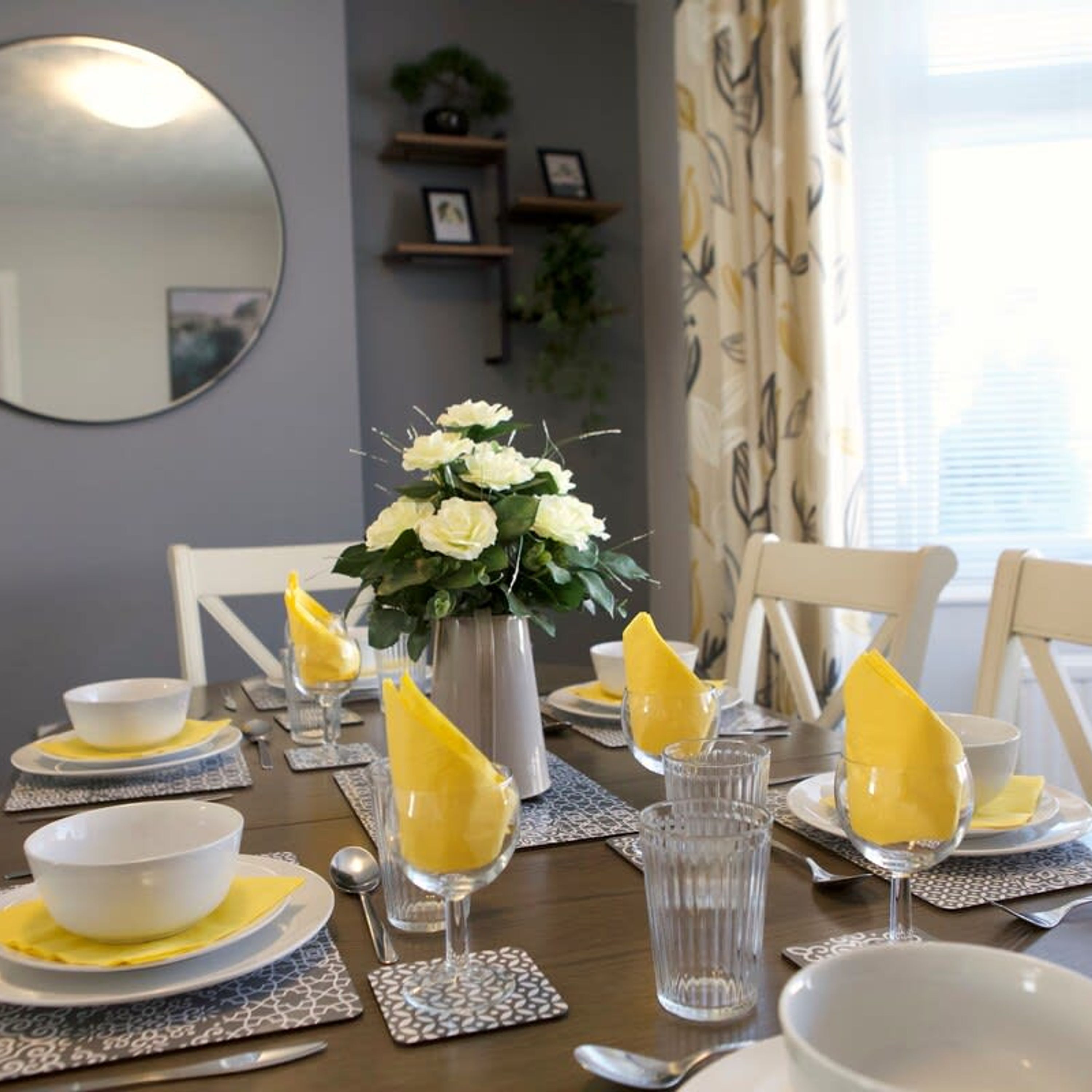 Una mesa de comedor dispuesta con servilletas amarillas, platos blancos y vasos transparentes.