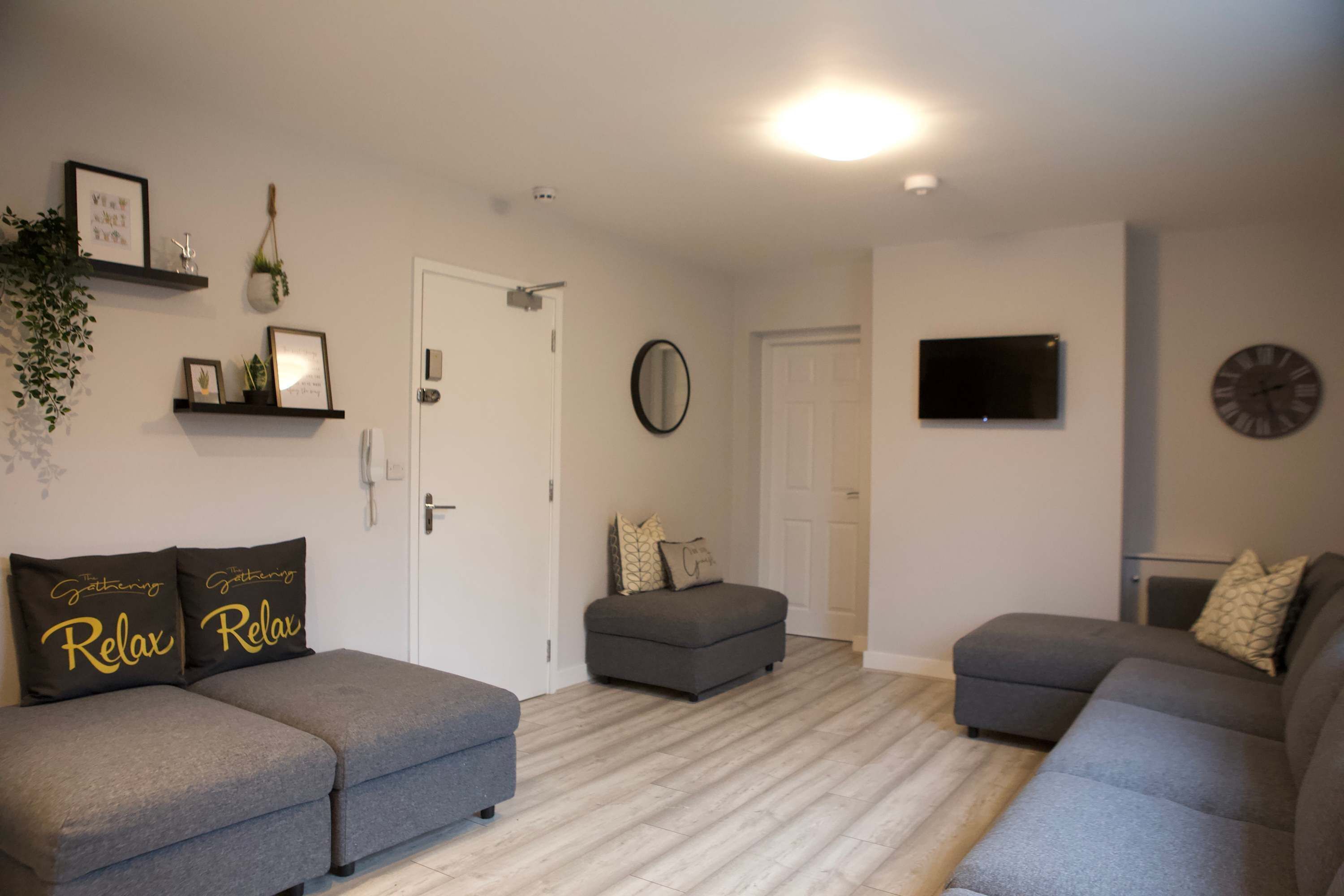 A living room with grey sofas, a grey ottoman, a flat-screen TV, a clock, and a door.