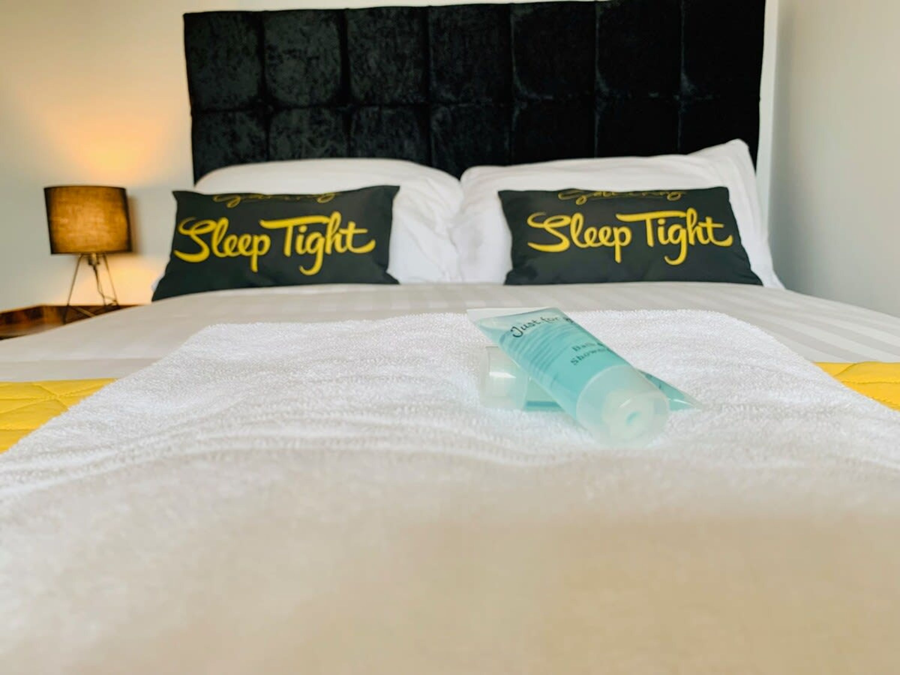 A neatly made bed with a white comforter and two pillows with 'Sleep Tight' text, a bedside lamp, and a bottle of lotion on the bed.