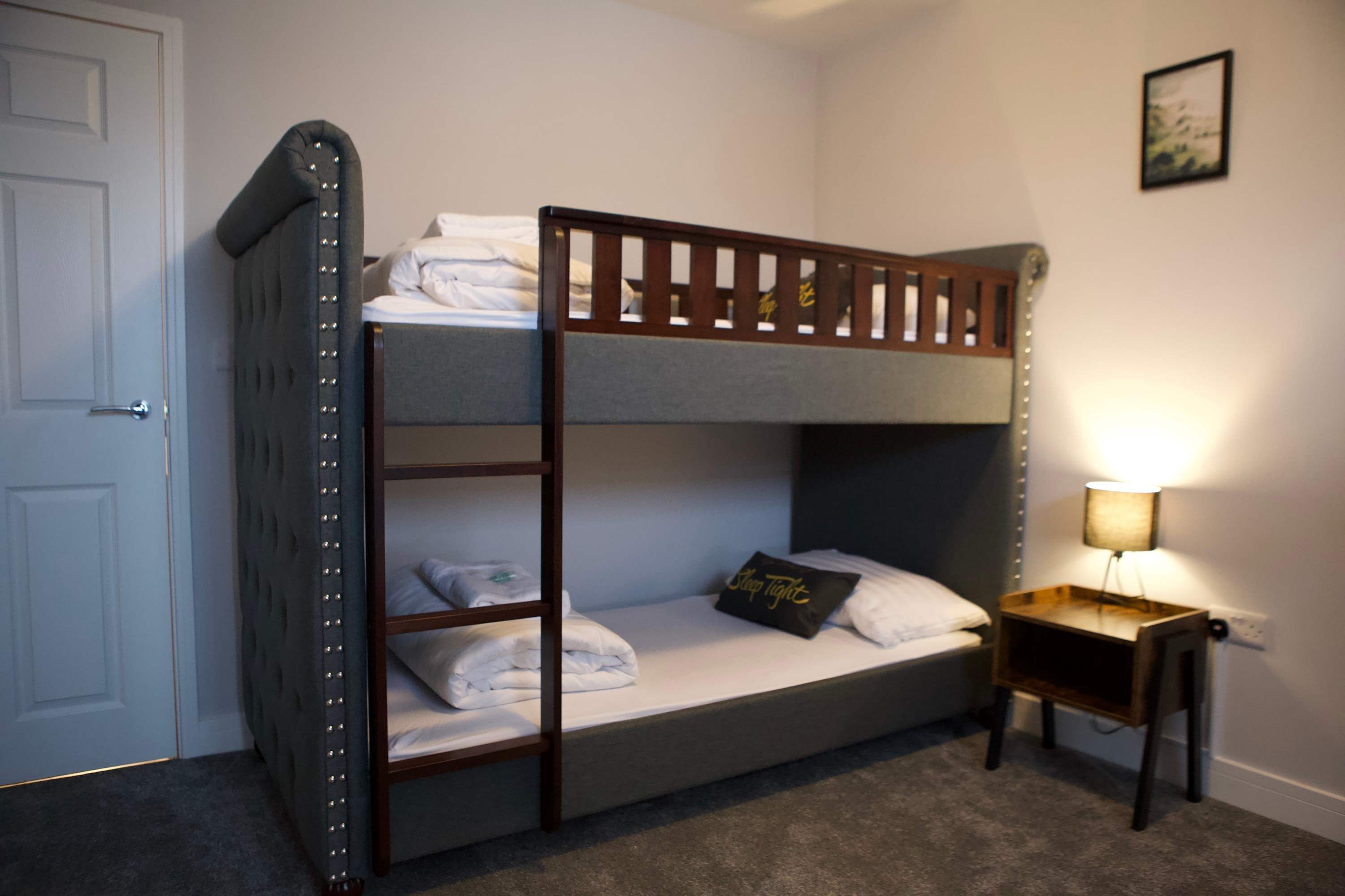 A bunk bed with a ladder, a bedside table with a lamp, and a door.