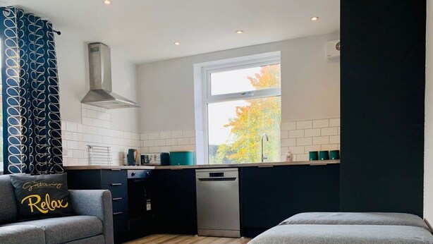 A modern kitchen with a dishwasher, a window with a view of autumn trees, and a grey sofa with a 'Relax' pillow. - The Gathering Sale - Sleeps Up To 22 (Sale)