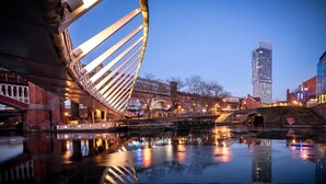 A modern bridge with a unique cable design, a river reflecting lights, and a high-rise building in the background. - The Islington Studio The Gathering Sale (Sale)