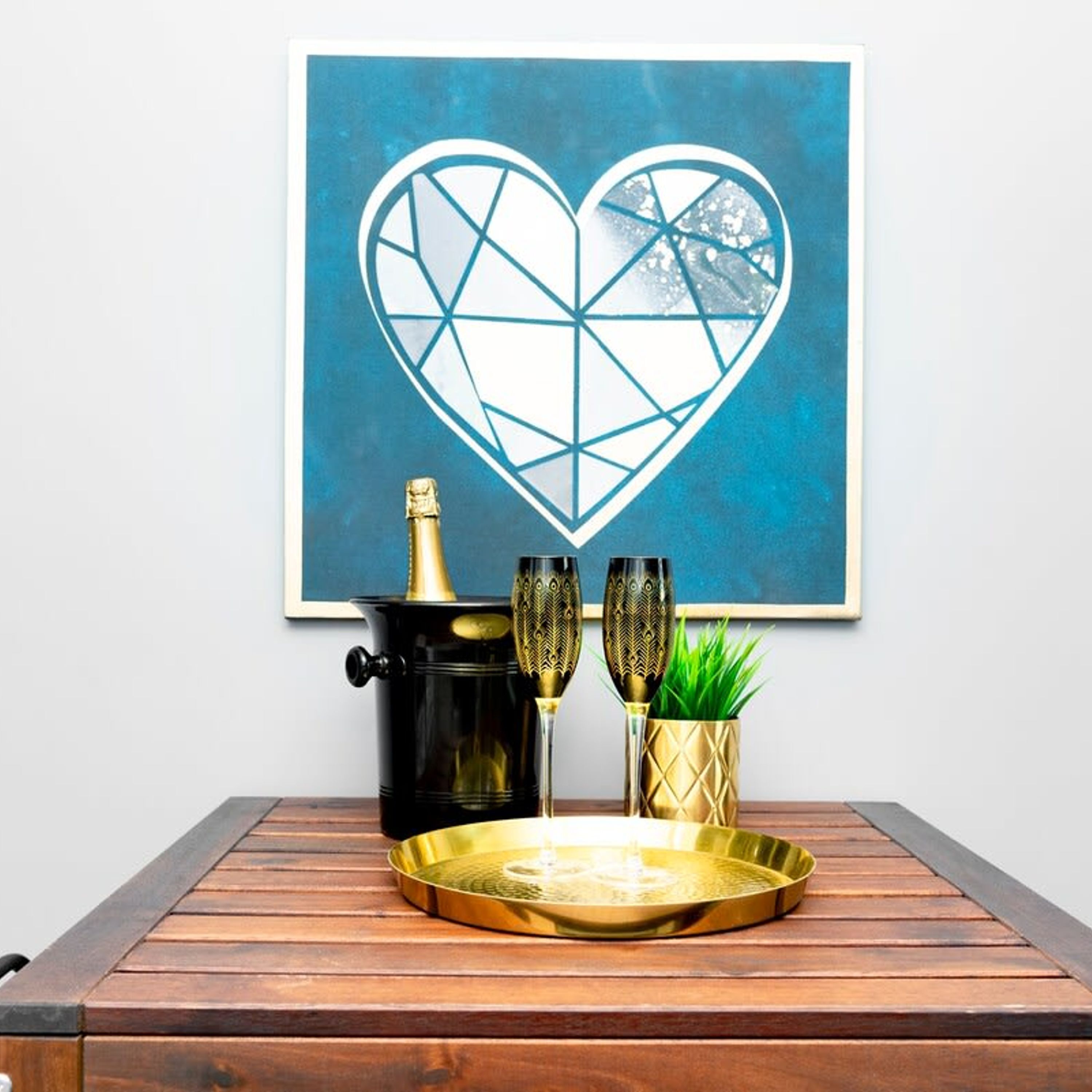 A framed abstract heart artwork on a wall, a black pot with a gold lid, two ornate glasses, a gold tray, and a potted plant on a wooden table.