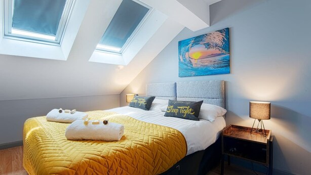 Quarto com cama, duas claraboias, um quadro, luminária de cabeceira e mesa de apoio.
